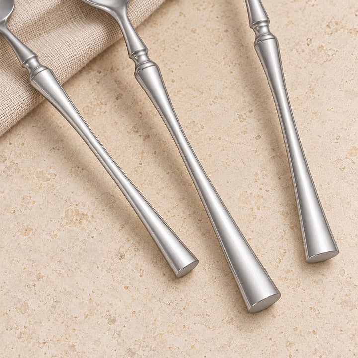 Three silver metal cutlery handles with elongated tapered handles arranged diagonally on a beige stone-like surface next to a folded beige fabric napkin.