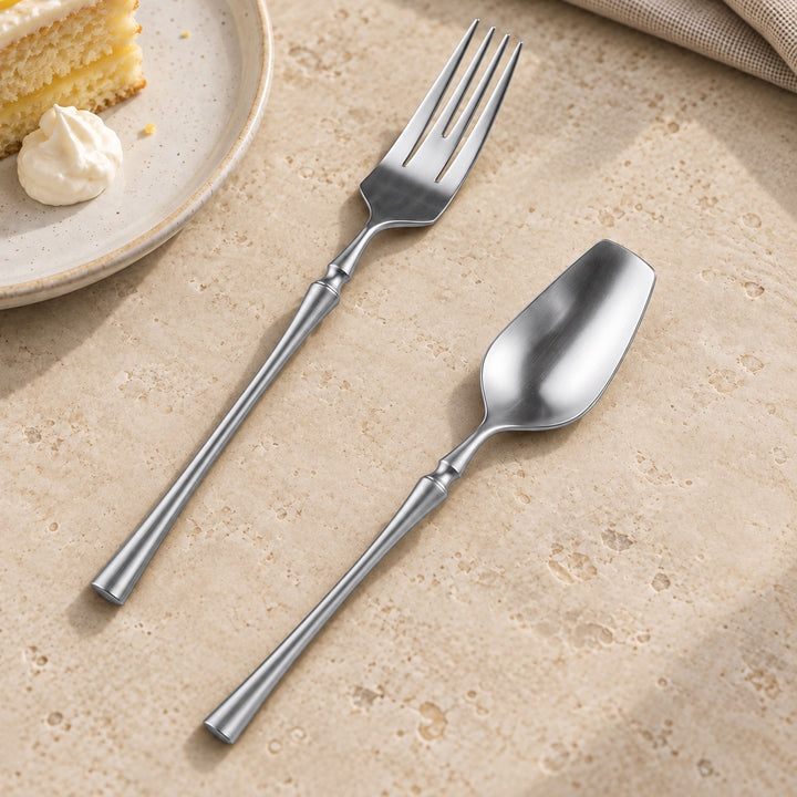 A stainless steel dinner fork and spoon with matching handles, arranged diagonally on a textured beige stone surface next to a plate with a slice of cake.