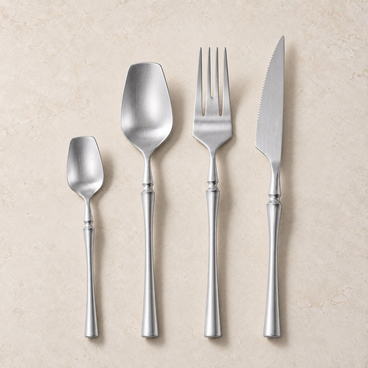 Four-piece stainless steel cutlery set with elegant handles, including a teaspoon, soup spoon, dinner fork, and dinner knife, arranged vertically on a textured beige background.