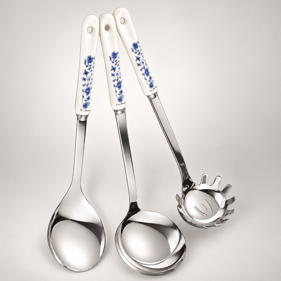 Three stainless steel kitchen utensils with white ceramic handles decorated with a blue floral pattern, arranged on a light grey background.
