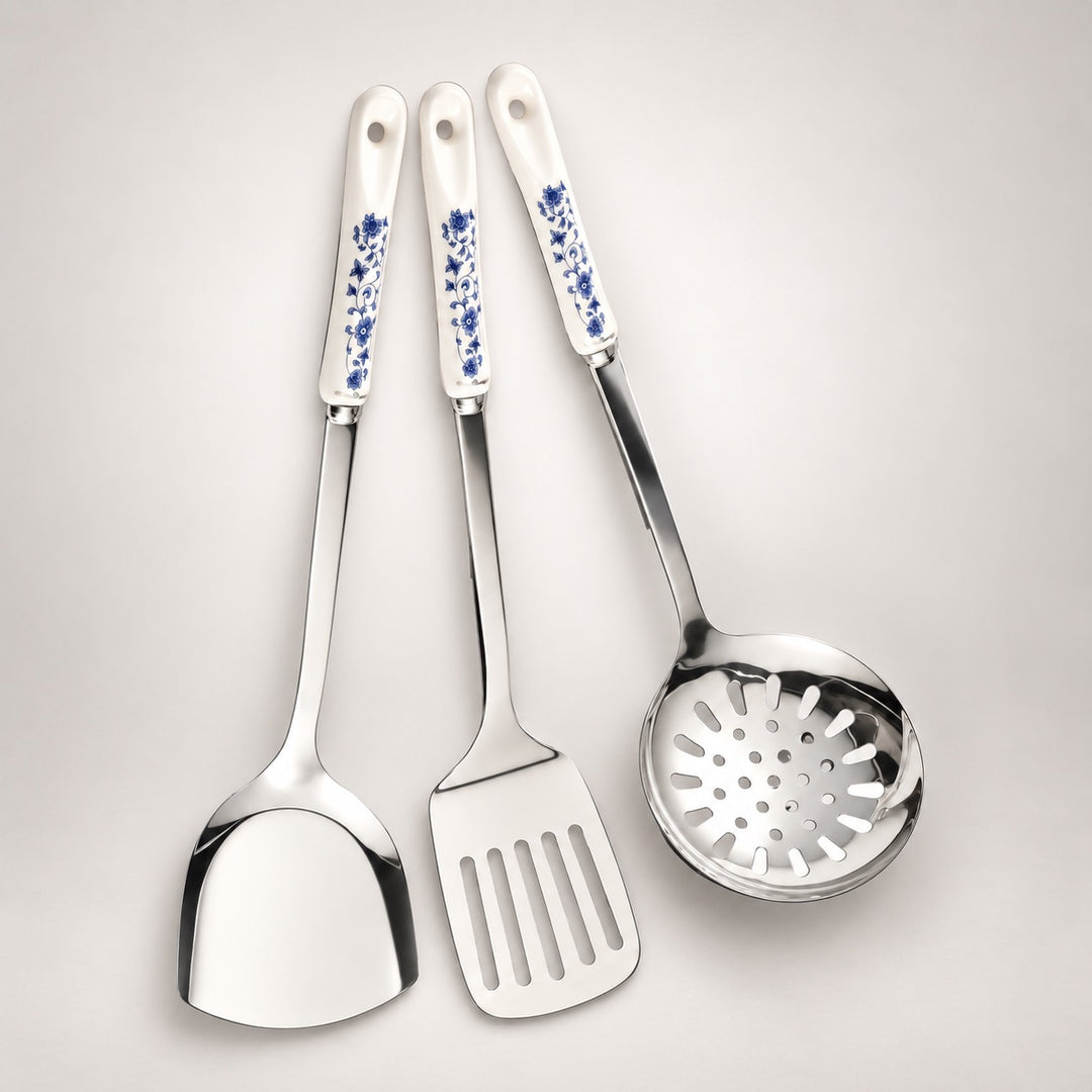 Three stainless steel kitchen utensils with white ceramic handles decorated with blue floral patterns, arranged on a light grey background.