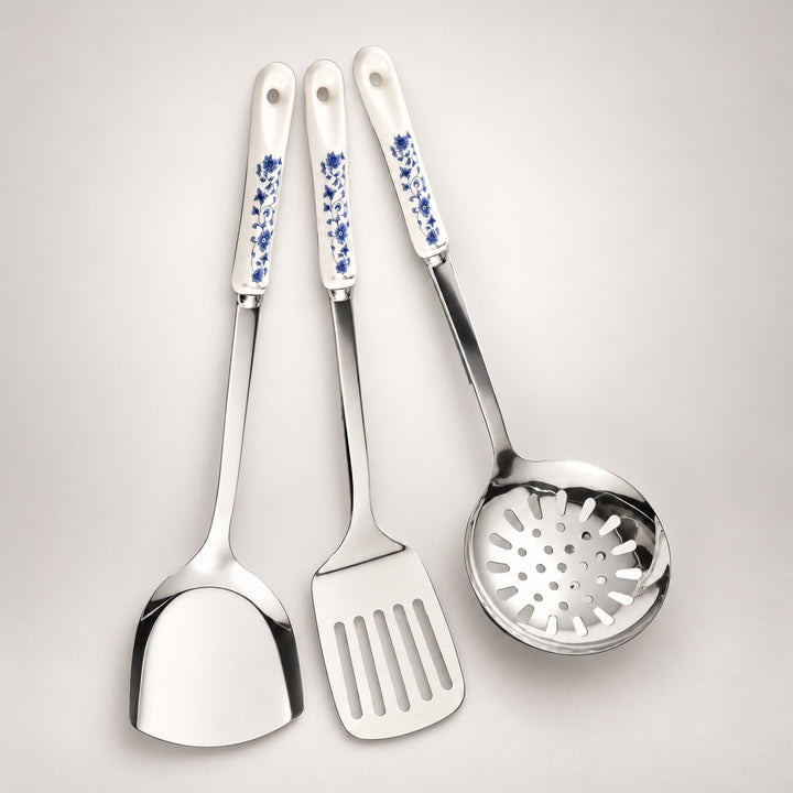 Three stainless steel kitchen utensils with white ceramic handles decorated with blue floral patterns, arranged on a light grey background.