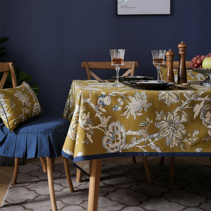 Dining table setting with floral-patterned tablecloth, chairs, plates, glasses, and fruit bowl.