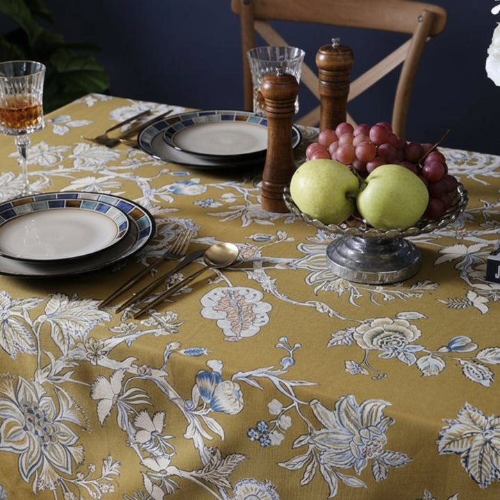 Dining table setting with floral-patterned tablecloth, plates, glasses, and fruit bowl.