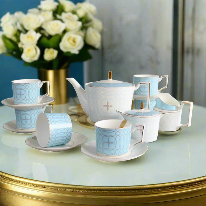 Elegant blue and white bone china tea set with gold accents on a glass and gold table, featuring a teapot, cups, saucers, a sugar bowl, and a milk jug, with a bouquet of white roses in the background.