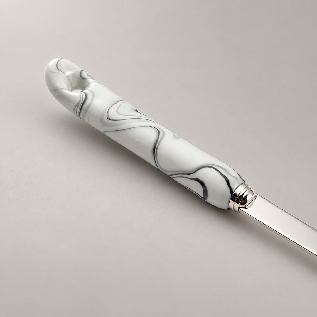 A close-up of a utensil with a grey ceramic handle featuring a black marble swirl pattern and a polished silver stainless steel, set against a neutral light grey background.