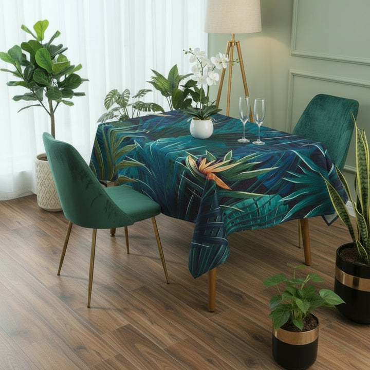 Dining table with a tropical leaf patterned tablecloth, surrounded by green chairs and potted plants.