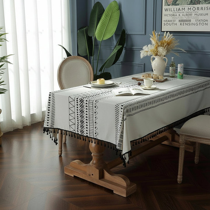 Dining room with a wooden table and white tablecloth featuring black patterns, surrounded by chairs and plants.