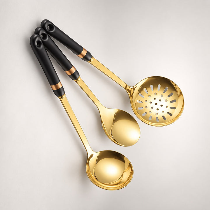 Three gold kitchen utensils with black handles and gold bands, including a ladle, spoon and slotted spoon, arranged diagonally on a light gray background.