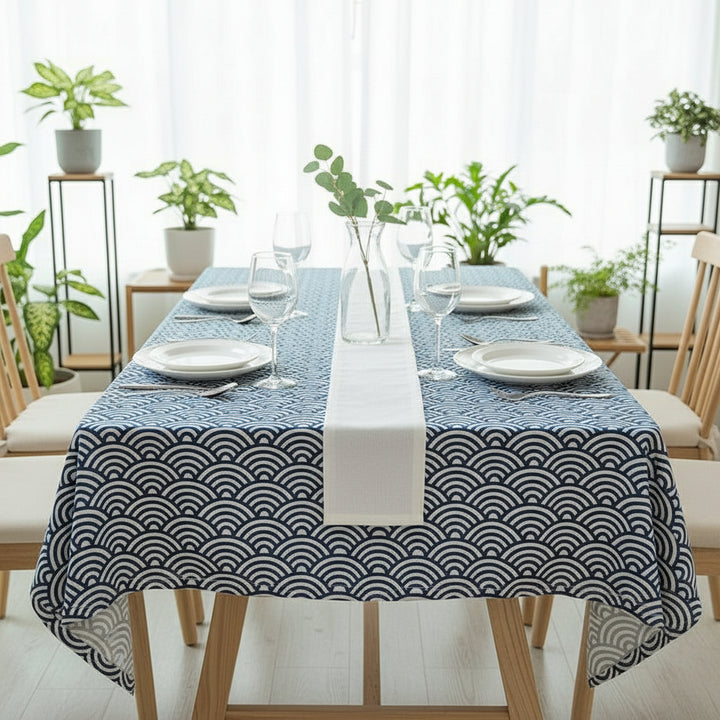 Dining table set with a blue and white patterned tablecloth in a bright room with plants.