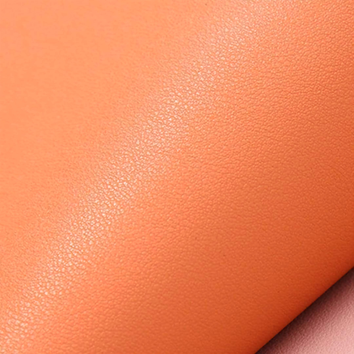 A close-up of a warm orange faux-leather placemat material with a soft diagonal fold showing the texture and colour.