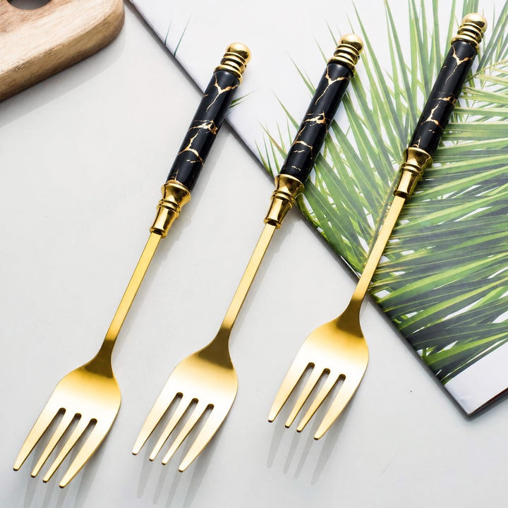 Three gold-plated salad forks with black marble-patterned handles, arranged diagonally on a light gray surface with a green palm leaf and a wooden board as props.