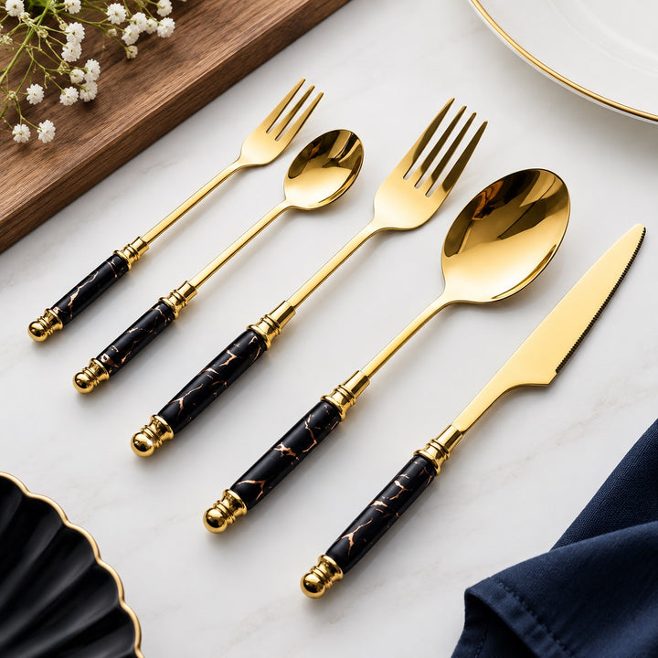 Five-piece gold-plated cutlery set with black marble-patterned handles, arranged diagonally on a white marble surface with decorative props.