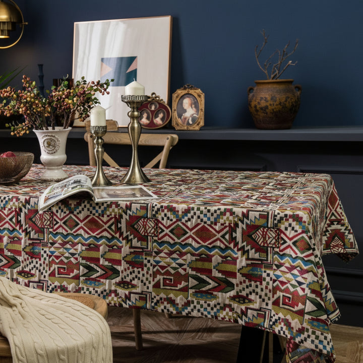 Decorative table setting with a patterned tablecloth, candles, and vases against a dark wall.